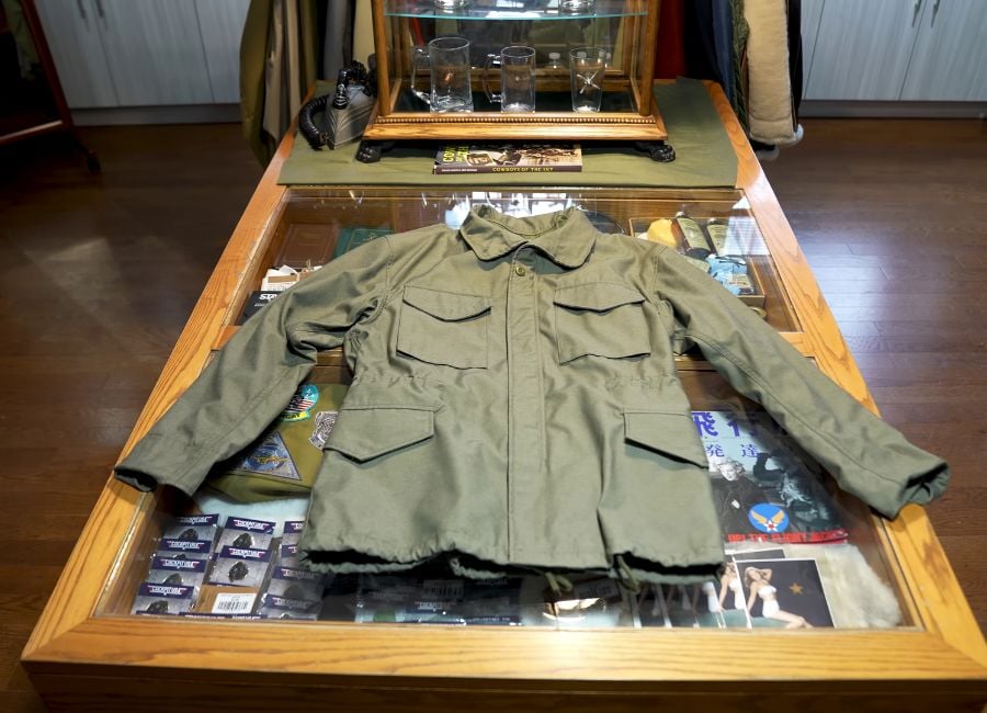 An army green M-65 Field Jacket by Cockpit USA laid out on a glass display case