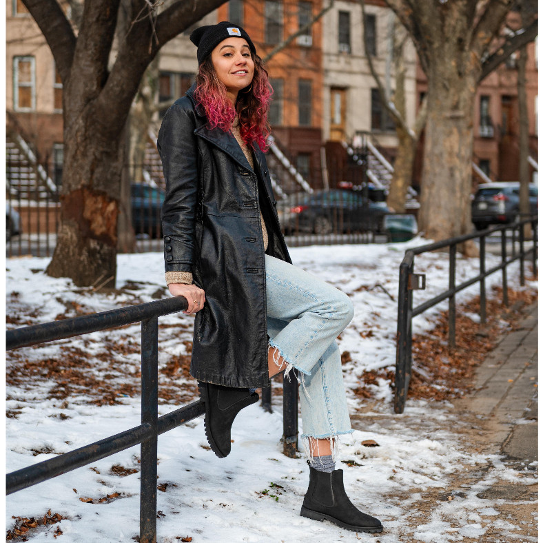 Hilary sitting on a rail with Thursday's women's legend chelsea boot in matte black wearing a leather jacket and light washed denim